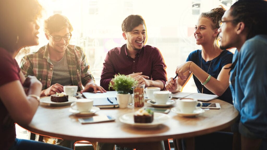 A small group of people having a relaxed conversation around a table in a café, suggesting trust, familiarity, and human connection.