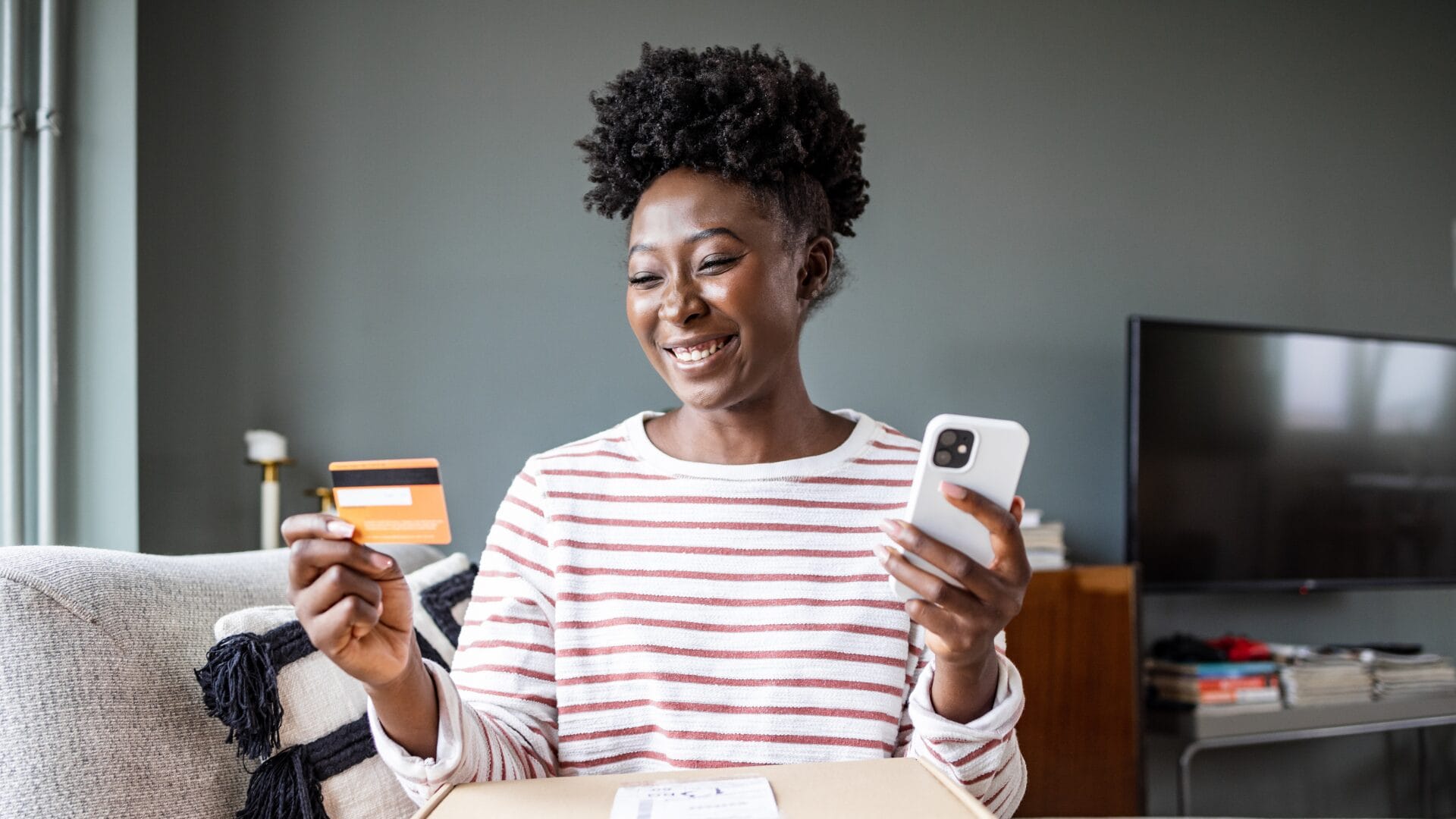 A smiling woman sitting on a couch holds a credit card and a smartphone while unboxing a package. The image represents a positive online shopping experience, highlighting trust, convenience, and seamless ecommerce transactions.