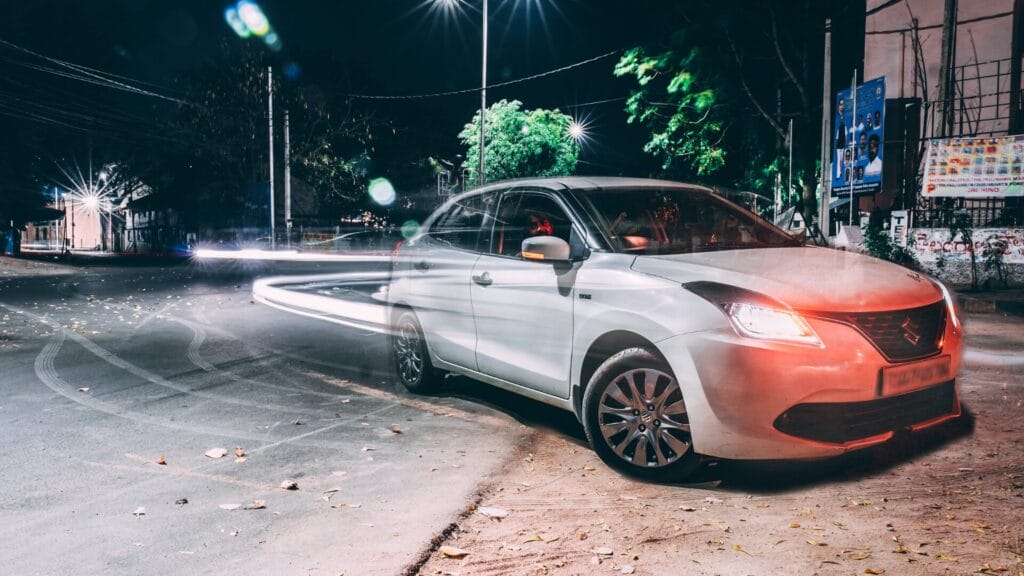 A white Suzuki car parked on a city street at night, surrounded by streetlights and vibrant light trails, reflecting a modern and urban ambiance.