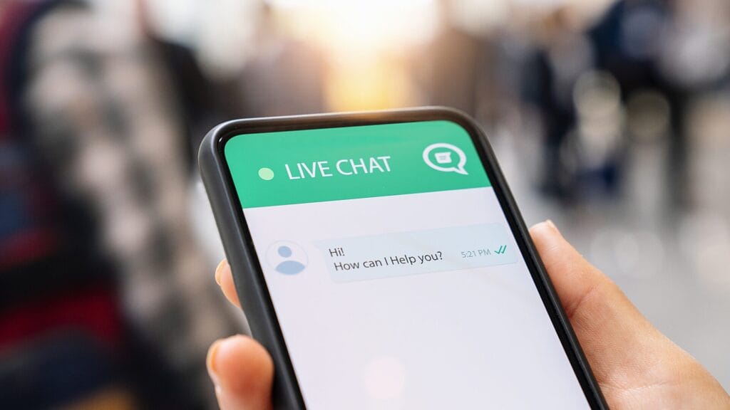 A hand holding a smartphone showing a live chat interface with a support message that reads, “Hi! How can I help you?” The background is softly blurred, suggesting a public or office environment.